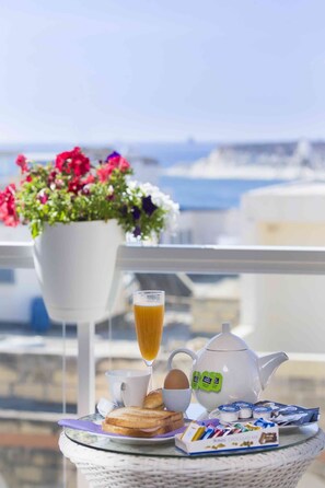 Enjoy a serene seaside breakfast on this inviting balcony with a wicker table, featuring a teapot, cup, and a selection of sweet and savory treats.