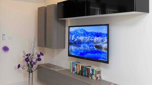 En moderne stue med et fladskærms-tv på væggen, et gråt tv-møbel nedenunder, og en vase med lilla blomster til venstre.