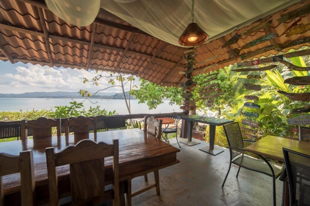 A covered outdoor dining area with wooden tables and chairs, overlooking a body of water.