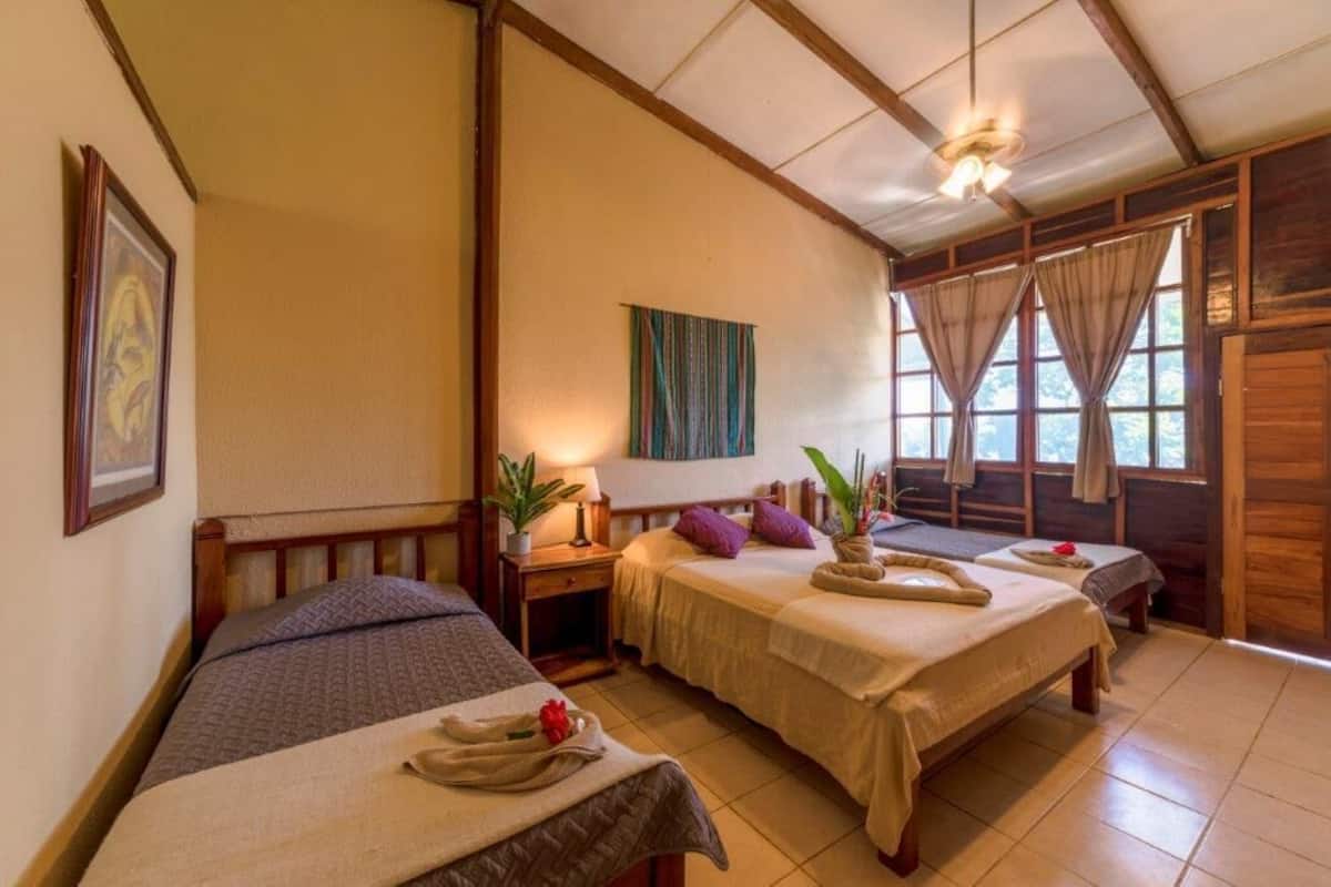 A room with two beds, a wooden ceiling, a wall-mounted painting, a window with curtains, and a small table with a lamp and a plant.
