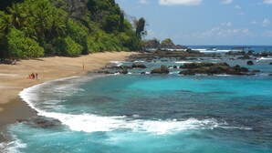 Uma praia com águas turquesa cristalinas, afloramentos rochosos e vegetação verde exuberante.