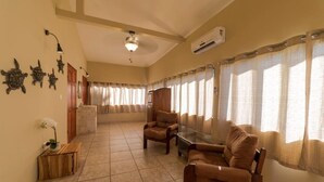 Ampla sala de estar com pé direito alto, ventilador de teto e lareira. Há duas poltronas de couro e uma mesa de centro de vidro. O ambiente possui grandes janelas com cortinas leves e vista para o exterior.
