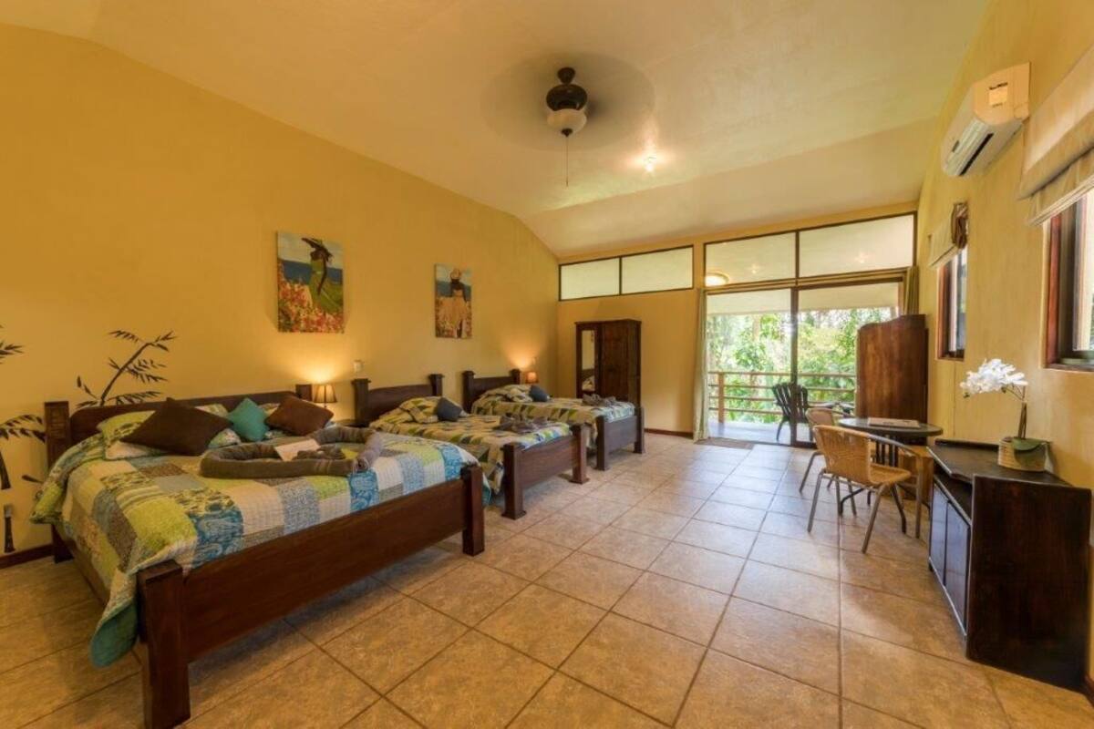 A room with three beds, a desk, and a chair. There are large windows and a ceiling fan.