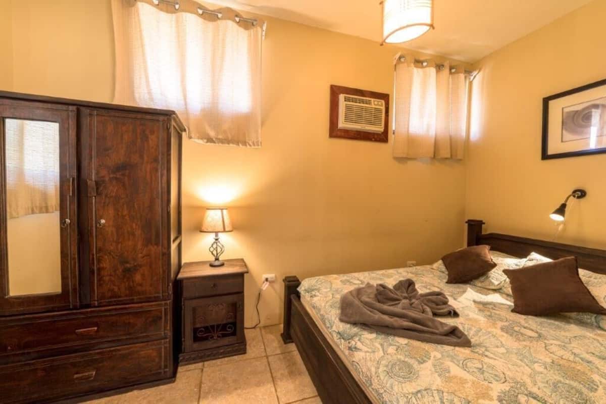 A bedroom with a wooden wardrobe, a bedside table with a lamp, a bed with patterned bedding and pillows, and a wall-mounted air conditioner.