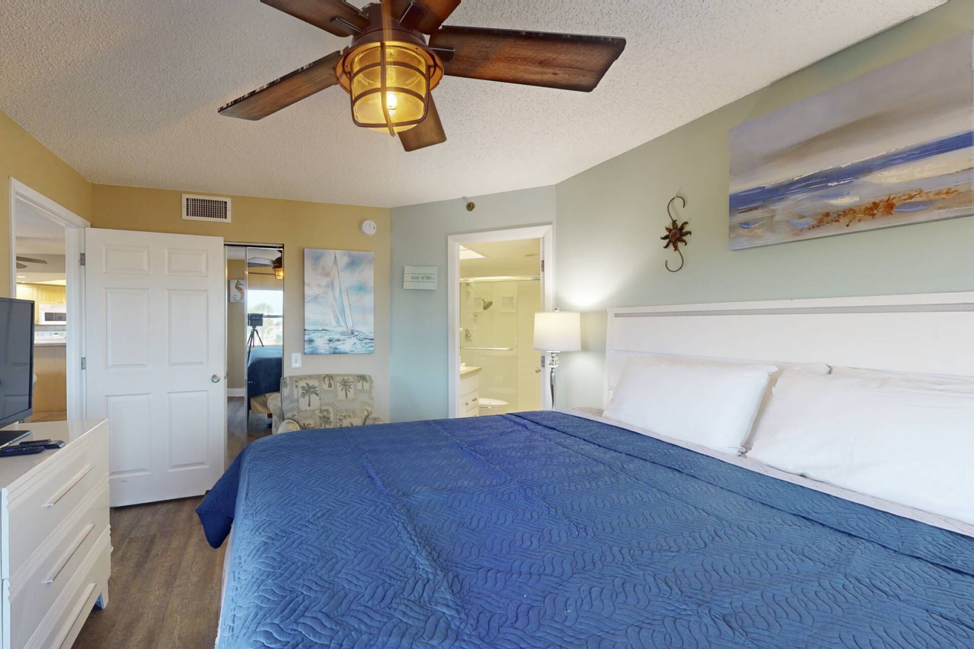 A bedroom with a bed, a ceiling fan, a painting, and a view of the bathroom.