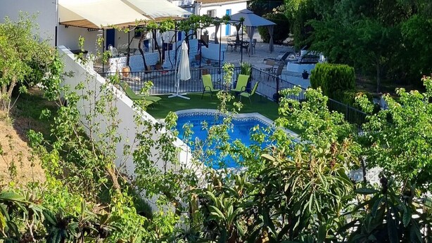 A pool area with lounge chairs and umbrellas, surrounded by greenery and a hillside in the background.