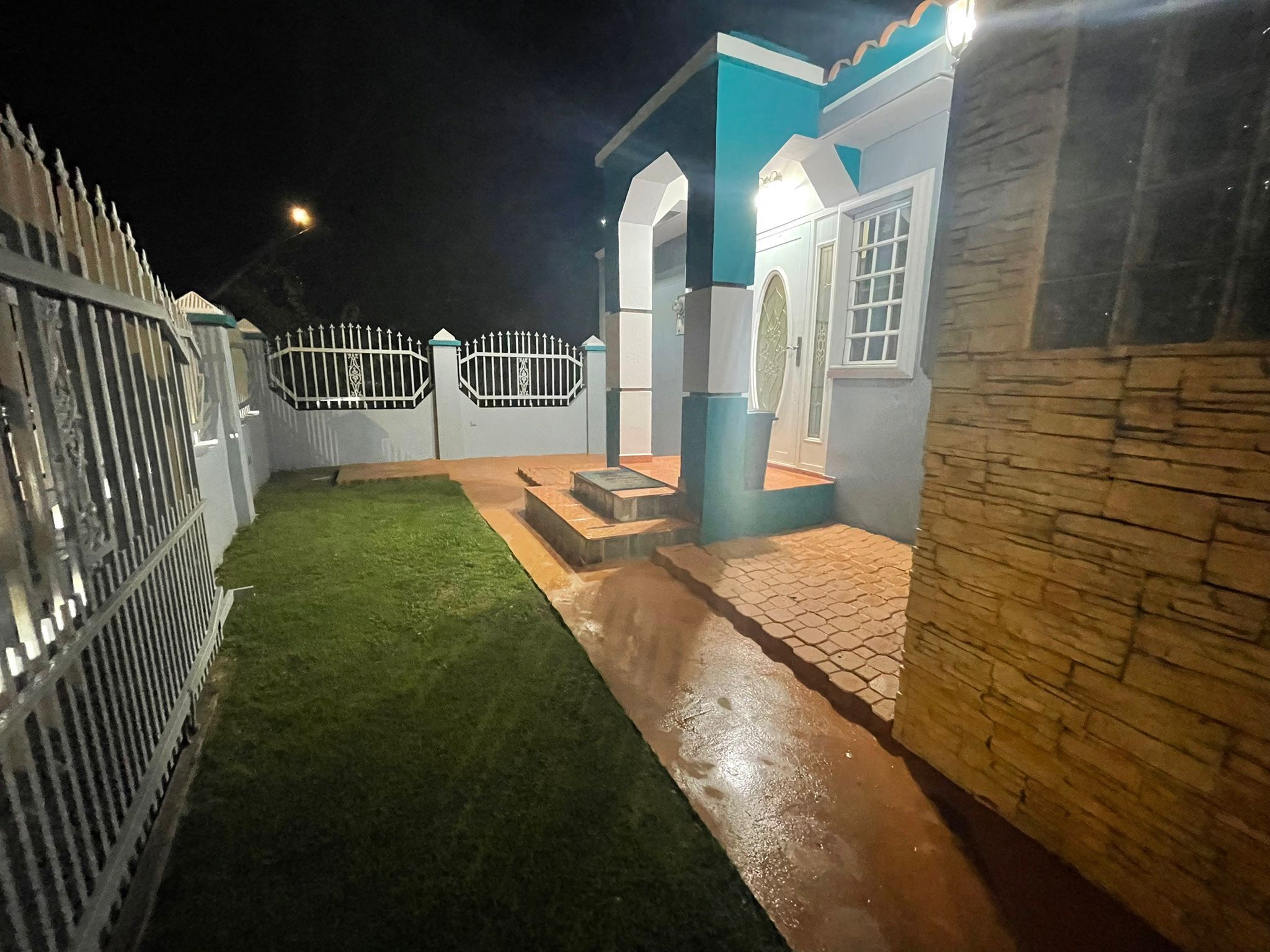A well-lit outdoor area with a paved walkway, a small grassy lawn, and a white fence.