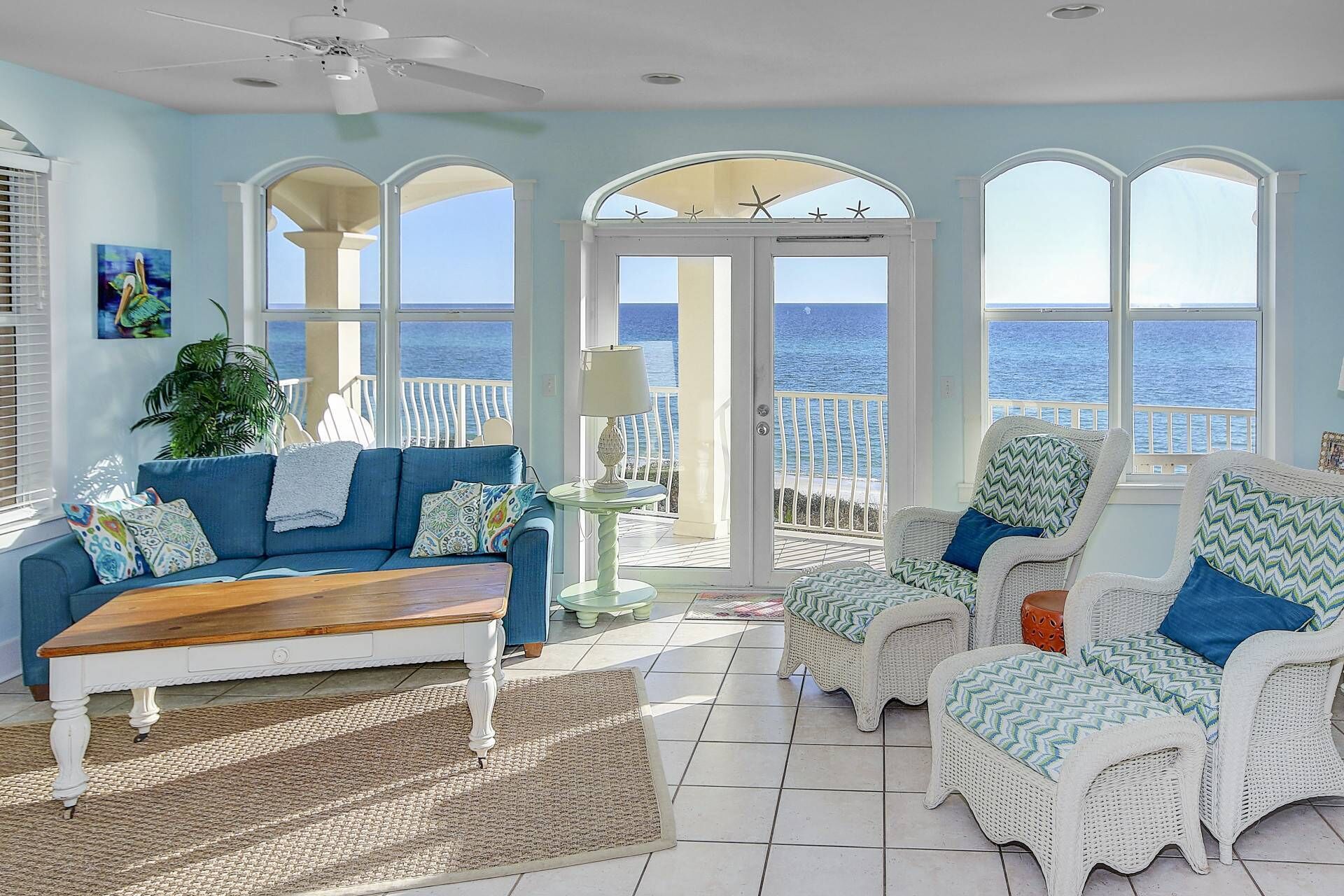 A coastal living room with a blue sofa, wicker chairs, a wooden coffee table, and a view of the ocean through large windows.