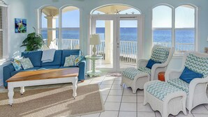 Un salón con vistas al mar, con sofá azul, sillas de mimbre, una mesa de centro de madera y grandes ventanales.