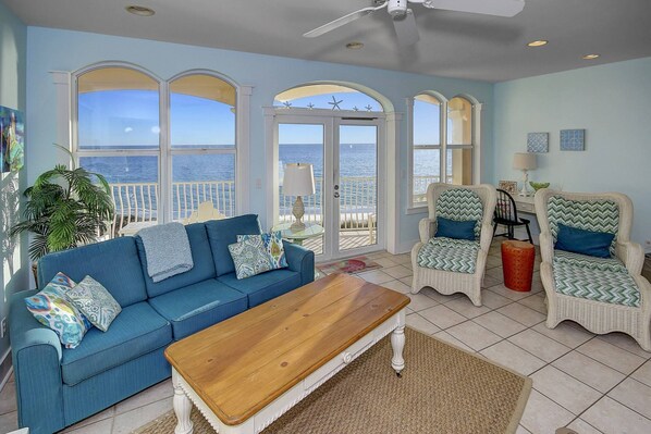 A spacious living room with a blue sofa, two wicker chairs, a wooden coffee table, and large windows offering a view of the ocean.