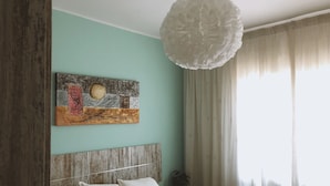 Un dormitorio con cabecera de madera, cama con ropa de cama blanca y marrón, lámpara colgante, un cuadro en la pared y cortinas de encaje en la ventana.