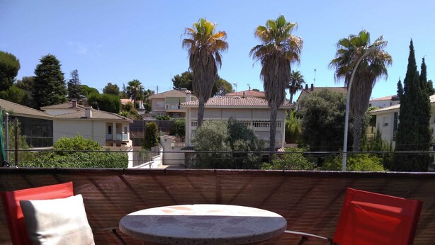 Un balcón con dos sillas rojas y una mesa redonda, con vista a una zona residencial con casas y palmeras.