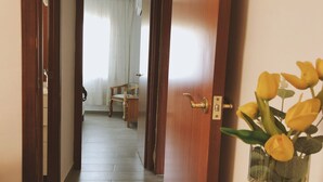 Puerta de madera con manija de latón que da a un cuarto con piso de madera, una silla y una mesa. A la derecha de la puerta, se ve un jarrón con flores amarillas.