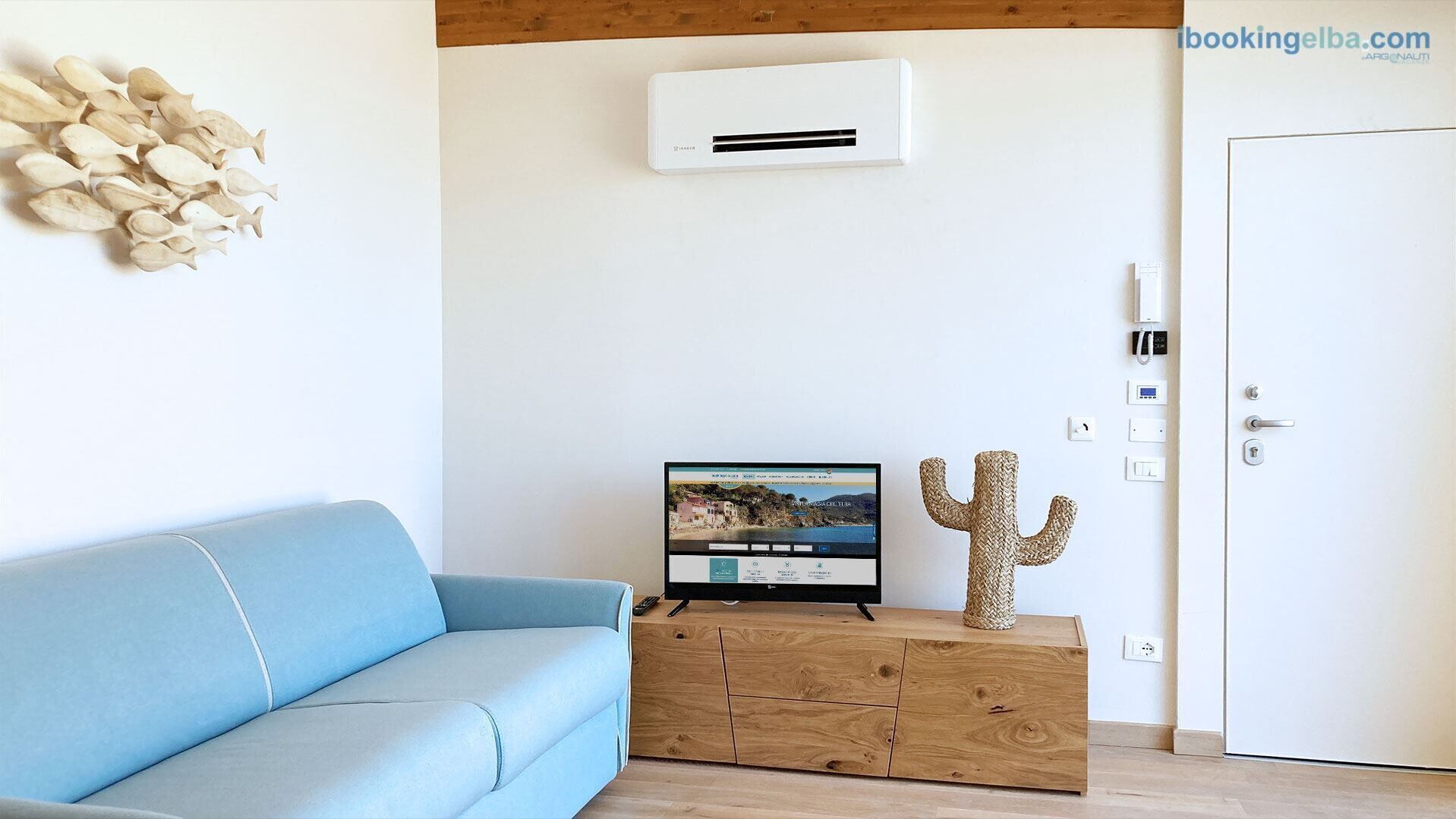 Une pièce avec un canapé bleu clair, un meuble en bois orné d’un motif de cactus, un climatiseur fixé au mur et un téléviseur affichant une image de paysage.