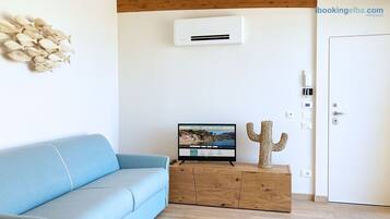 Une pièce avec un canapé bleu clair, un meuble en bois orné d’un motif de cactus, un climatiseur fixé au mur et un téléviseur affichant une image de paysage.