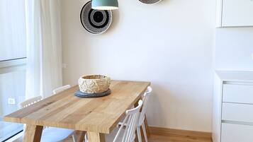 Une table de salle à manger en bois, agrémentée d’un centre de table en forme de panier tressé, des chaises blanches, un luminaire fixé au mur et un meuble blanc.