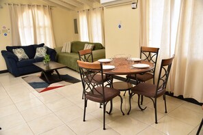 Ein Essbereich mit einem runden Tisch, vier Stühlen und einem Couchtisch. Im Hintergrund befindet sich eine Couch und ein grüner Sessel.