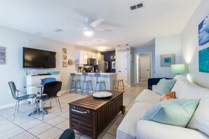 A modern living room with a TV, a coffee table, a sofa, and a kitchen area in the background. - Grand Beach 207 | Pool + Hot Tub + Gulf Views (Gulf Shores)
