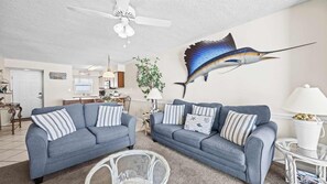 A living room with two grey sofas, a ceiling fan, a dining area with a table and chairs, and a wall-mounted fish decoration.