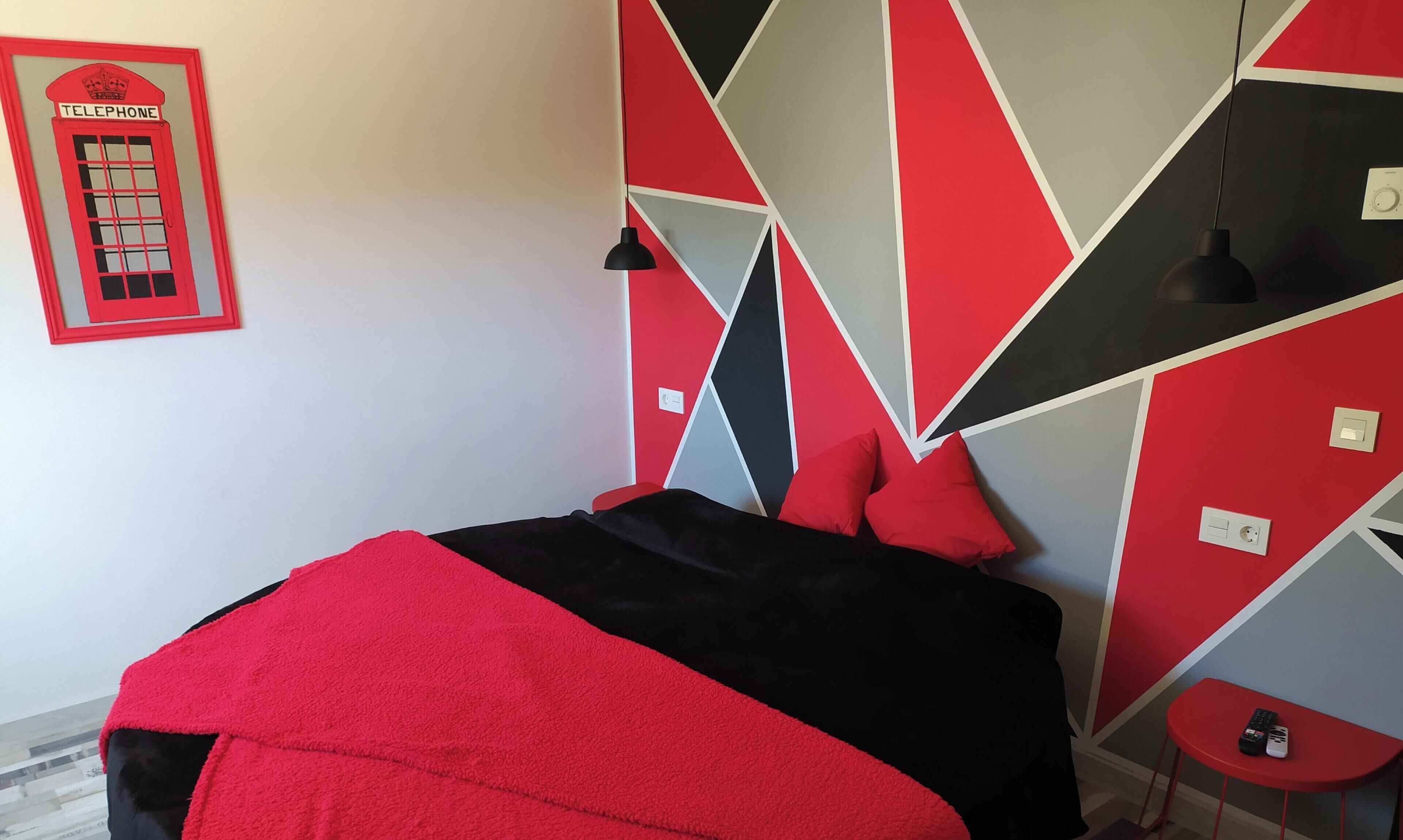 A modern bedroom with a geometric-patterned wall, a bed with red and black bedding, a red side table, and a wall-mounted telephone.