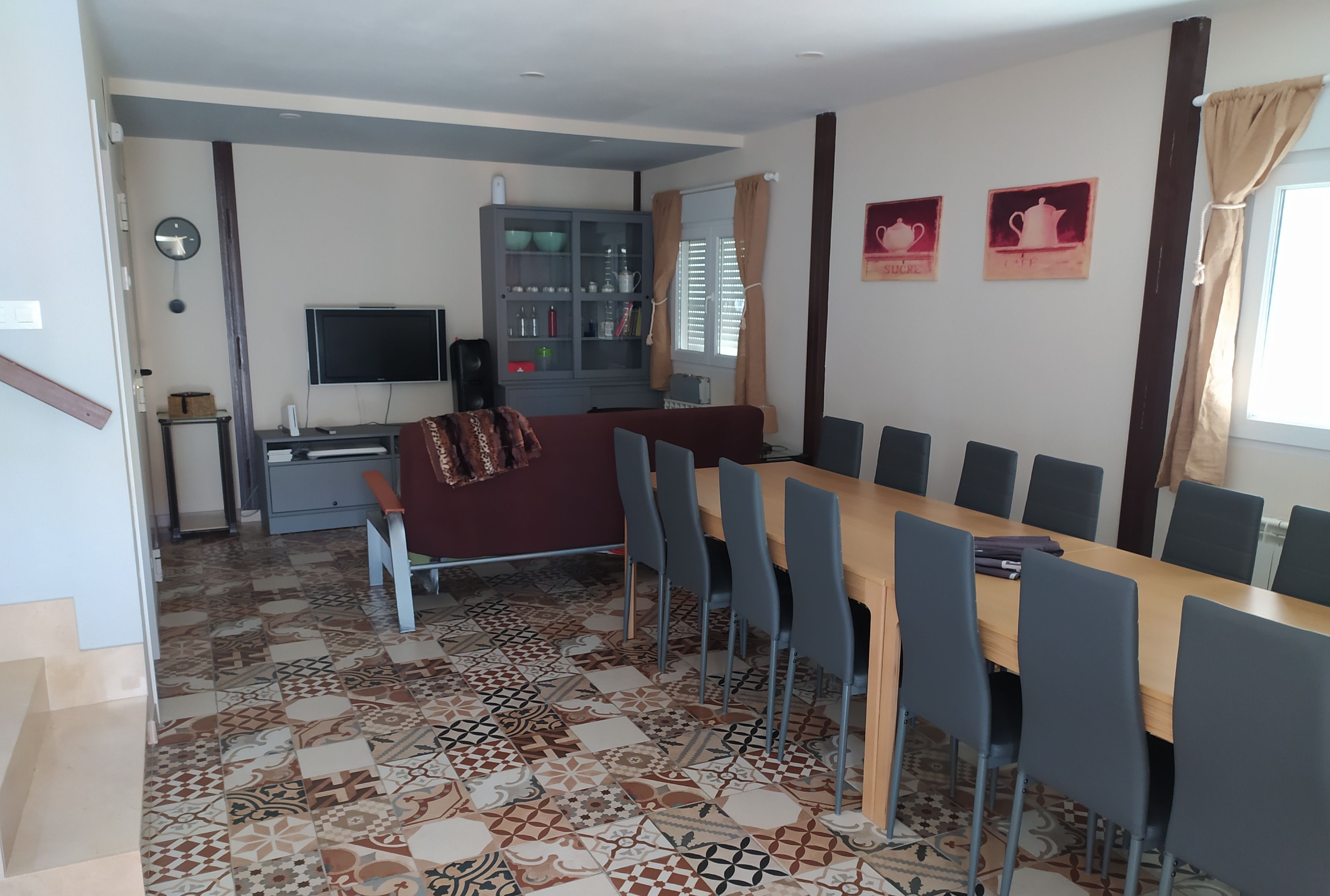Een eetgedeelte met een lange tafel en stoelen, een televisie, een kast met glazen deuren en vloertegels met een patroon.
