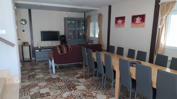 Een eetgedeelte met een lange tafel en stoelen, een televisie, een kast met glazen deuren en vloertegels met een patroon.