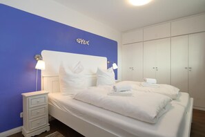 A bedroom with a bed, white nightstands, a blue accent wall, and a wardrobe. - At Undine Beach, Germany (Sylt)