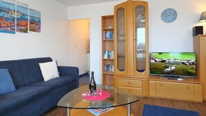 Een woonkamer met een blauwe bank, een glazen salontafel met een fles en glazen, een houten kast met glazen deuren, een televisie en een wandklok.
