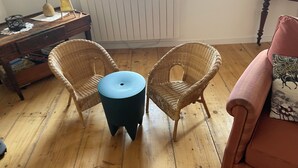 Een kamer met twee rieten stoelen, een kleine blauwe tafel, een roze bank, een houten vloer, een radiator en een raam met jaloezieën.