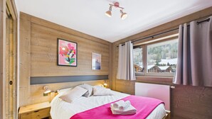 A wooden bedroom with a bed, a nightstand, a window with curtains, and a view of a snowy landscape. - Cozy apartment in the heart of the village with terrace and garage - Les Gets (Les Gets)