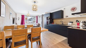 A modern kitchen with black cabinets, a wooden dining table, and a view of a snowy landscape through large windows. - Cozy apartment in the heart of the village with terrace and garage - Les Gets (Les Gets)
