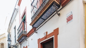 Une rue étroite avec un trottoir à carreaux, un immeuble avec une porte marron et un balcon, et une pancarte indiquant qu’un bien est à vendre.