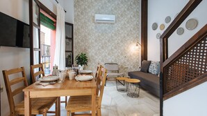 Un coin repas avec une table en bois, des chaises et un canapé. Un climatiseur est fixé au mur, et des assiettes décoratives sont accrochées au mur.