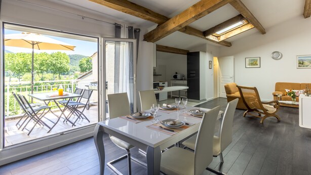 A dining area with a table set for four, a kitchen in the background, and a view of the outdoors through large windows. - "l'escapade"... Entre Lacs, mer et Montagnes (Hérépian)