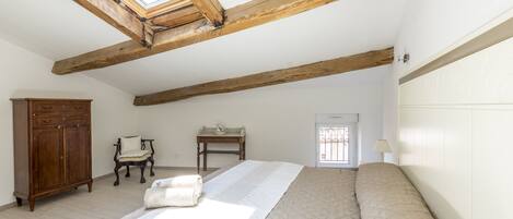 A bedroom with a large bed, wooden beams, a wardrobe, a chair, a small table, and a window.