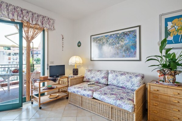 A living room with a sofa, TV, lamp, wicker furniture, and artwork. - GuestHost - Comfortable 50 sqm apartment with terrace, able to accommodate up to 4 people, located on the fifth floor of a building with lift (NOT suitable for disabled people - 15 steps to access it). The property is located in a quiet area of Tao (Taormina)
