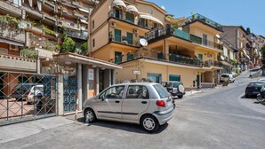 A residential street with multi-story buildings, parked cars, and a small car parked in front of a building with a gated entrance. - GuestHost - Comfortable 50 sqm apartment with terrace, able to accommodate up to 4 people, located on the fifth floor of a building with lift (NOT suitable for disabled people - 15 steps to access it). The property is located in a quiet area of ​​Tao (Taormina)
