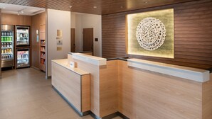 A reception area with a wooden reception desk, a wall-mounted decorative piece, and a refrigerator stocked with beverages. - Fairfield Inn & Suites by Marriott Memphis Arlington (Arlington)