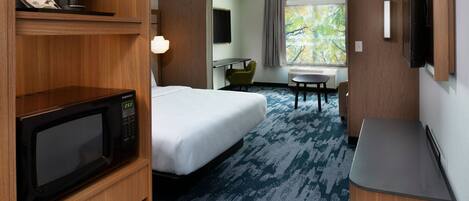 A hotel room with a bed, a microwave, a desk, a chair, a TV, and a window with a view of trees.