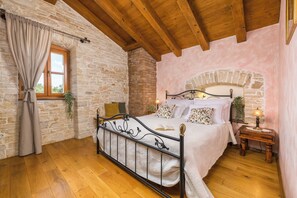 Ein Schlafzimmer mit Steinwand, Holzbalkendecke, einem Bett mit Metallgestell, einem Ziegelkamin und einem kleinen Nachttisch.