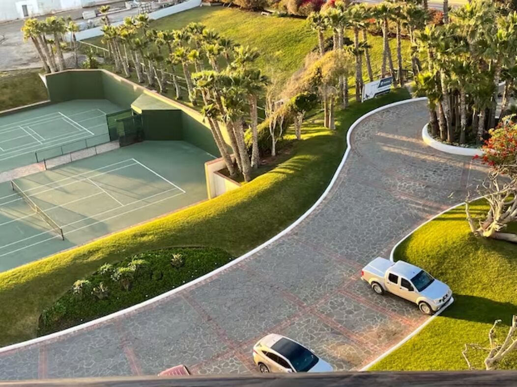 Luftaufnahme eines Tennisplatzes, umgeben von Palmen und einer gewundenen Auffahrt mit zwei geparkten Autos.
