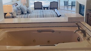 A bedroom with a large bathtub, a bed, a ceiling fan, and a view of the ocean.
