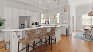 A modern kitchen with white cabinetry, a stainless steel refrigerator, a large island with bar stools, and pendant lighting. - Satilla Cottage - 4 Bedrooms in Coast Cottages (Saint Simons Island)