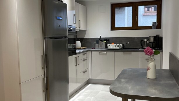 Una cocina moderna con un refrigerador de acero inoxidable, armarios blancos, una encimera gris y una pequeña mesa con un jarrón.