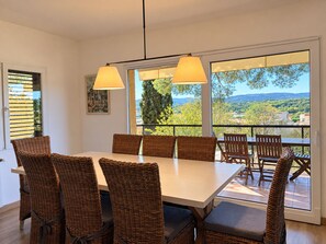 Un coin repas avec une grande table et des chaises en osier, de grandes fenêtres donnant sur un paysage verdoyant et des collines, et un tableau accroché au mur.