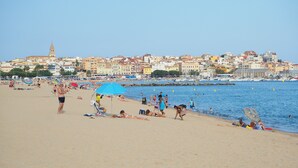 Una playa con gente tomando el sol y nadando, cielo despejado y edificios al fondo.