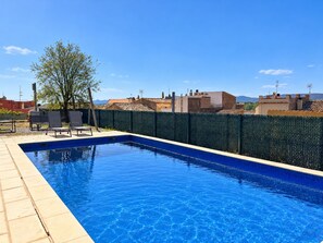 Une piscine rectangulaire bordée de carreaux, entourée d’une clôture verte, avec un ciel bleu clair en arrière-plan.