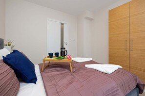 A bedroom with two beds, a wooden wardrobe, a bedside table with a coffee pot, cups, and a plant.