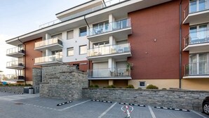 A multi-story residential building with balconies, stone pillars, and a paved parking area. - Apartment with Balcony | Pool, Gym, 100 m to the Beach (Rewal)