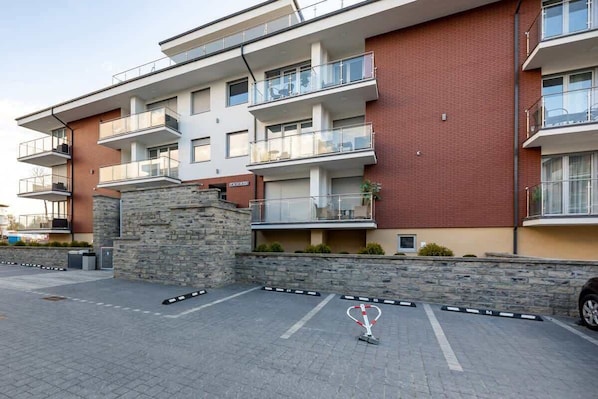 Un edificio residencial de varios pisos con balcones, pilares de piedra y un estacionamiento pavimentado.